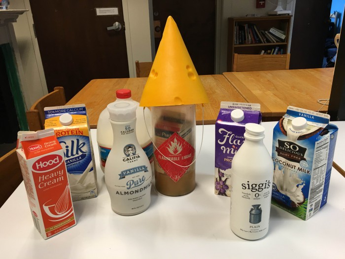 Photo of a container with a yellow cone party hat, surrounded by half a dozen containers of other varieties of milk, including almond, soy, coconut, and skyr