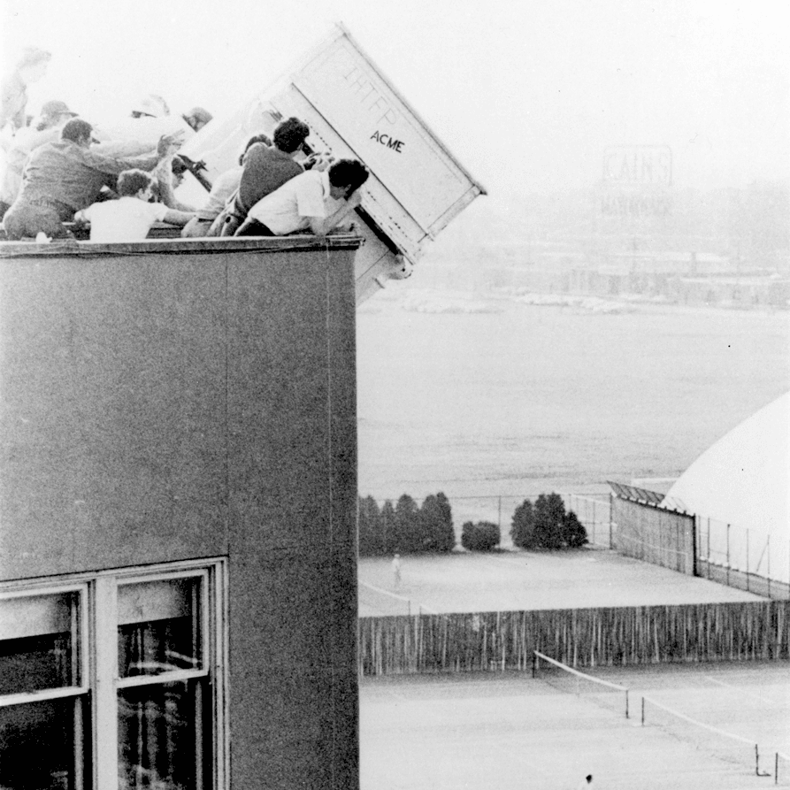 Scan of a black and white photo of students raising a piano about to drop over the edge of the building