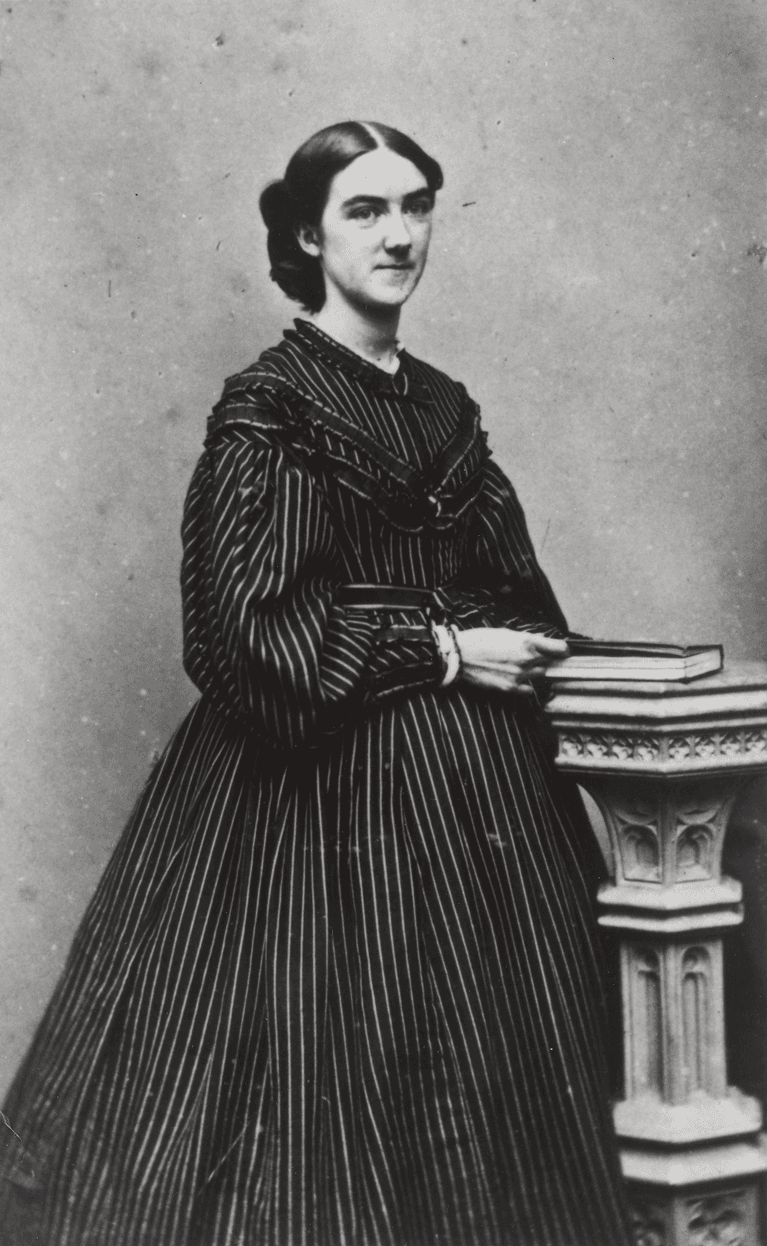 scan of black and white photo of a woman in striped victorian dress, standing next to a pillar shaped pedestal with a book on top.