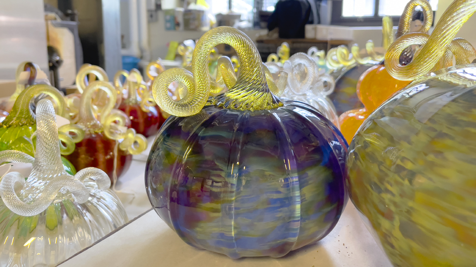 Photo of dozens of glass pumpkins, front and center is a purple/blue/multi-colored pumpkin