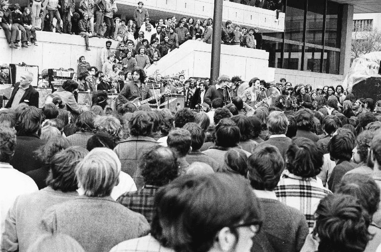 Black and white photo of Grateful Dead perform in front of the MIT Stratton Student Center with crowds standing on all around them including on the steps.